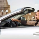 Homem dirigindo um carro conversível na frente do Coliseu de Roma.