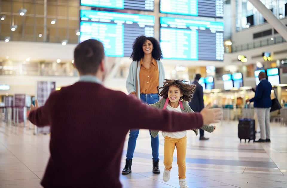Criança no aeroporto correndo em direção ao pai, com a mãe ao fundo.